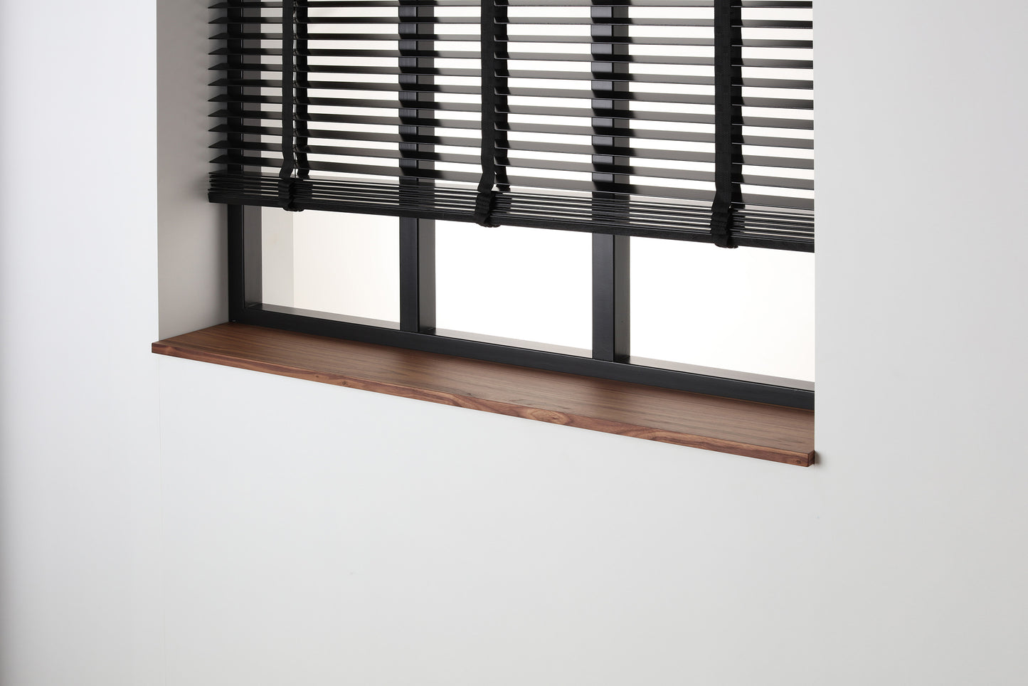 Noten gefineerde vensterbank met matte blanke lak onder een zwart raam met jaloezieën.