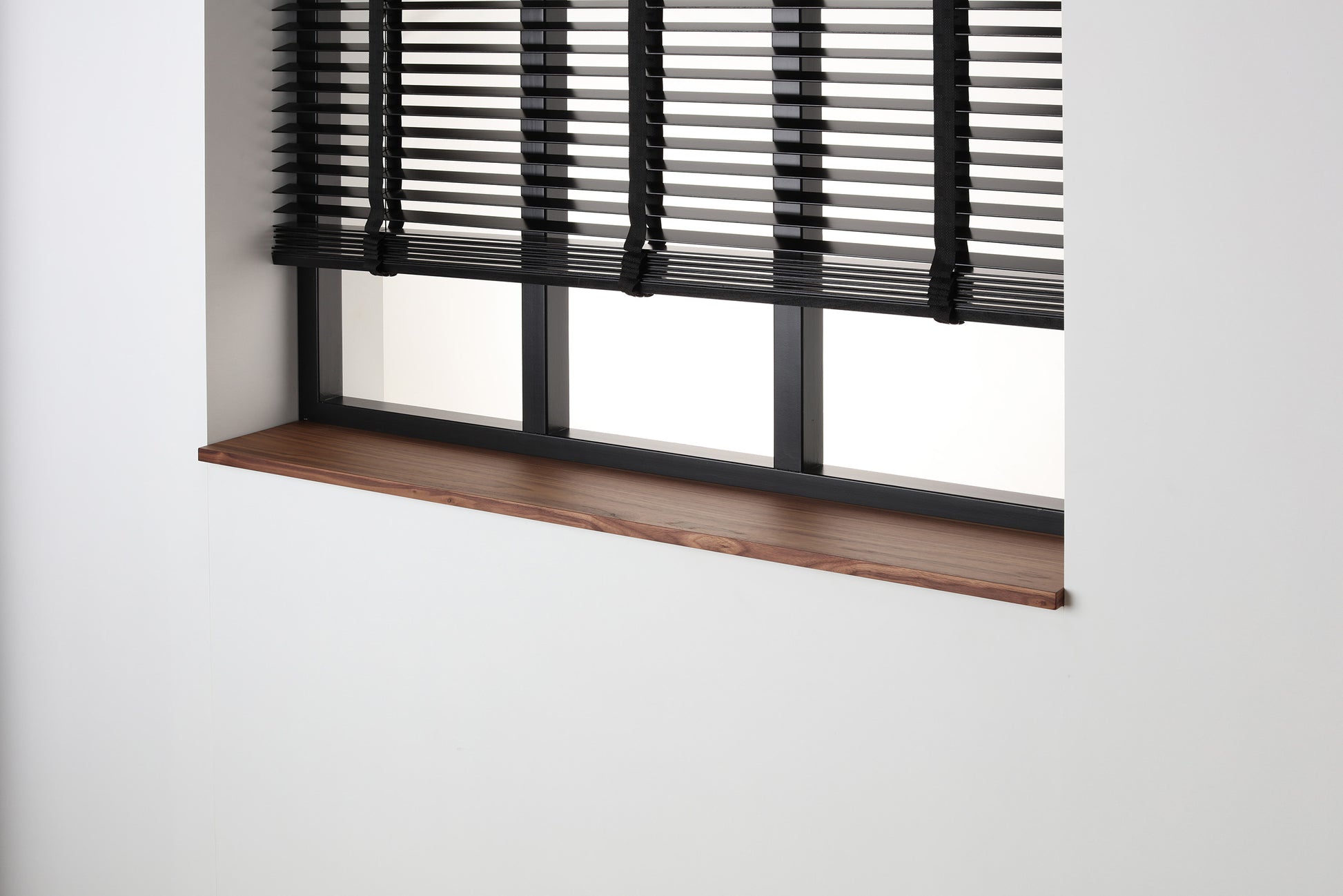 Noten gefineerde vensterbank met matte blanke lak onder een zwart raam met jaloezieën.